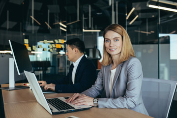 Portrait of successful woman, business team member, business woman looking at camera and smiling working on laptop in modern office