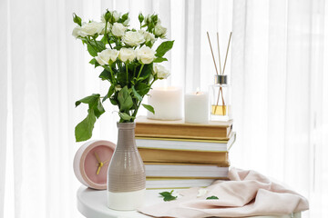 Vase with bouquet of beautiful roses, books, clock and reed diffuser on table in room