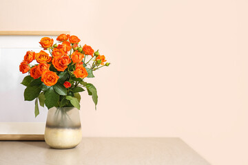 Vase with beautiful orange roses and blank photo frame on table against light wall