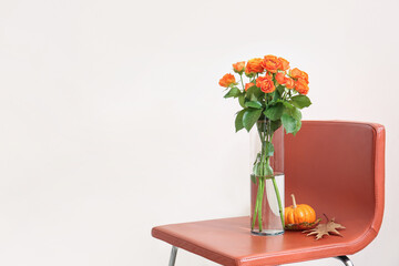 Vase with beautiful orange roses and pumpkin on chair against light wall