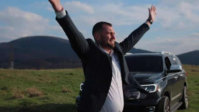 Businessman stands on a background of mountains near a black car raises his hands and shouts.