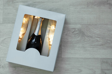 A bottle of champagne and two glasses with a luminous garland in a cardboard box. On white wooden background.