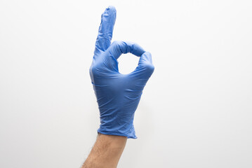 Mano con guantes azules de vinilo sobre fondo blanco, haciendo el gesto de bien con los dedos, de...
