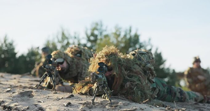 Military men in moro uniforms dressed in ghillie camouflage grass moss men lie on ground, guns on stand with scope prepare to fire on attacker from concealment training ground territorial defense