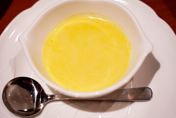 A stylish small white bowl of sweet corn soup made of brand corn from Hokkaido Japan with a round metal spoon and a white plate underneath