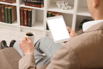 Man with tablet computer and cup of coffee sitting in armchair at home, closeup