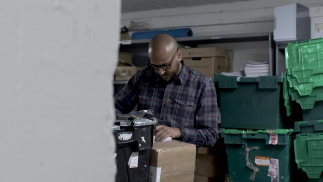 A static shot peaking in on an Indian man searching through boxes of old company documents in a cluttered storeroom, the man finding a card of interest setting it aside and continuing his search