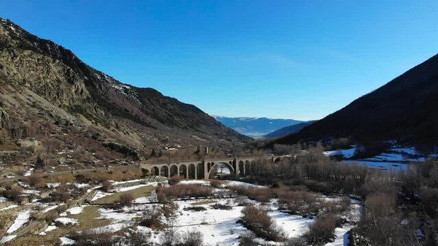 Aerial: magnificent stone bridge for a train track in the Pyrenees with some castle ruins in the backgraound and blues sky in a partly snowy lansdcape