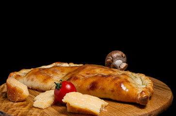 calzone closed pizza, on a wooden kitchen board, champignon mushroom, cherry tomatoes, parmesan cheese. Isolated on a black background.