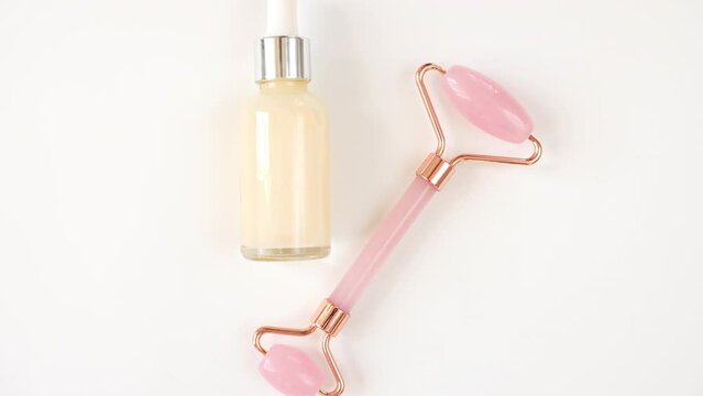 Rotation of pink quartz facial roller and glass bottle with gel, fluid isolated on white background, flat lay. High quality 4k footage