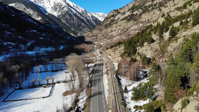 Aerial: sunny valley in the french Pyrenees with a road with little traffic, a train track and snowy mountains in the background
