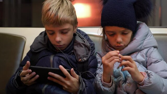 Children with headphones are watching videos on mobile phone on city underground train. Internet technology for streaming video on mobile phone in underground transport. Conscious use free time on way