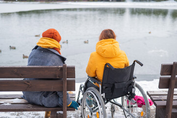 Caucasian woman in a wheelchair and her friend are sitting by the lake with ducks in winter.
