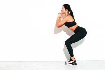 Fototapeta premium Portrait of fitness confident woman in black sports clothing. Sexy young beautiful model with perfect body. Female isolated on white wall in studio. Stretching out before training. Making squats