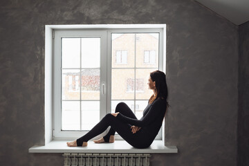 A woman in sportswear sits on the windowsill and looks out the window at the snowfall