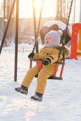 toddler 12-17 months old swings on a swing in winter on a children's playground. Sunbeam