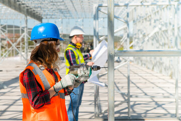 The professional young women worker works on installation of a metal frame using drill to screw and young engineer looking at the blueprint