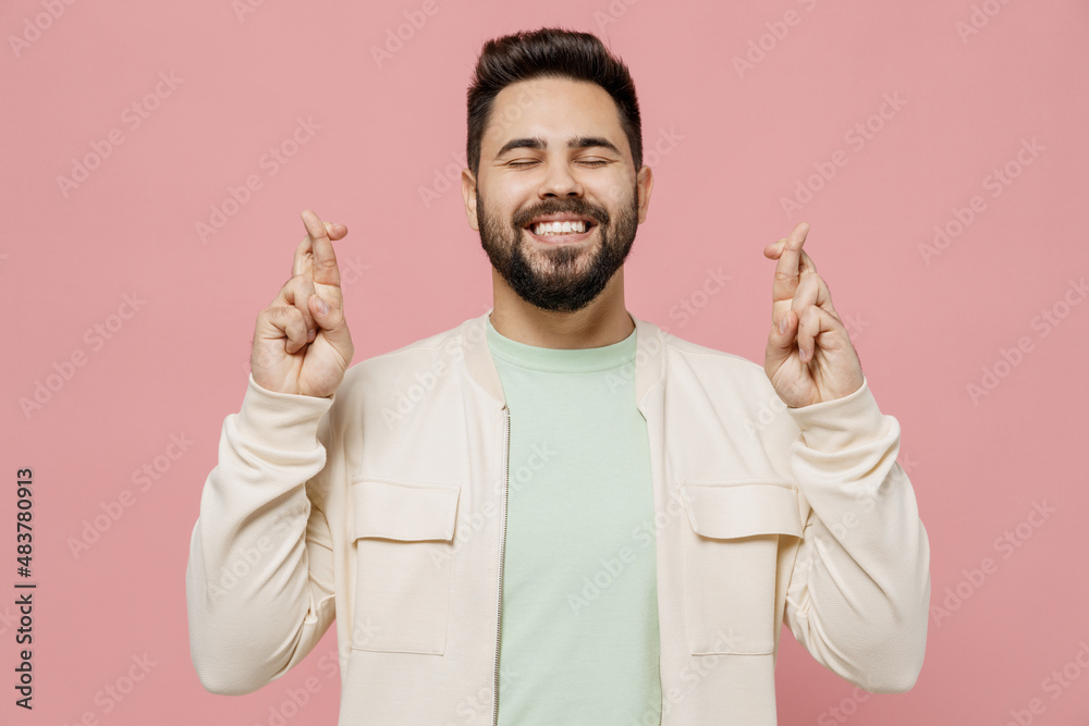 Wall mural Young man in trendy jacket shirt waiting for special moment, keeping fingers crossed, making wish, eyes closed isolated on plain pastel light pink background studio portrait. People lifestyle concept.