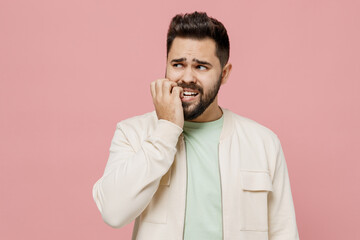 Young puzzled mistaken confused sad caucasian man 20s wear trendy jacket shirt look aside biting nails fingers isolated on plain pastel light pink background studio portrait. People lifestyle concept.