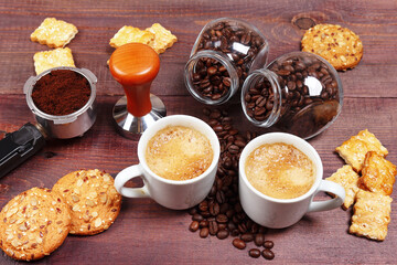 Two cups of coffee, crackers, holder with ground coffee, tamper and coffee beans on wooden table.