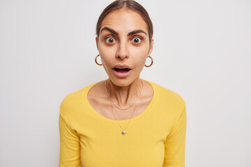 Amazed beautiful young woman with dark combed hair keeps mouth opened says wow and stares impressed at camera poses against white background drops jaw from big news. Human reactions concept.