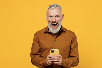 Shocked surprised happy elderly gray-haired bearded man 40s years old wears brown shirt look camera hold use mobile cell phone keep mouth wide open isolated on plain yellow background studio portrait.