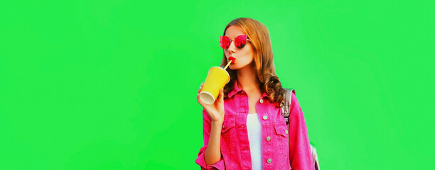 Portrait of stylish young woman drinking juice wearing pink jacket, sunglasses on green background