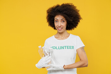 Young smiling woman of African American ethnicity wears white volunteer t-shirt gloves hold plastic pet bottle isolated on plain yellow background. Voluntary free work assistance help grace concept.