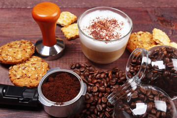 Breakfast with cappuccino, crackers, holder with ground coffee, tamper and coffee beans on wooden table.