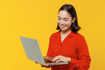 Businesslike smiling young woman of Asian ethnicity 20s years old wears orange shirt hold use work on laptop pc computer typing searching chatting isolated on plain yellow background studio portrait.