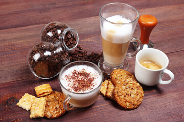 Breakfast with espresso, latte and cappuccino, crackers, holder with ground coffee, tamper and cans of coffee beans on wooden table.