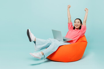 Full body young happy woman of Asian ethnicity 20s in pink sweater sit in bag chair use work on laptop pc computer with outstretched hands finish job isolated on pastel plain light blue background