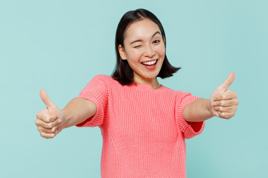 Young smiling fun happy woman of Asian ethnicity 20s wearing pink sweater showing thumb up like gesture blink isolated on pastel plain light blue background studio portrait. People lifestyle concept.