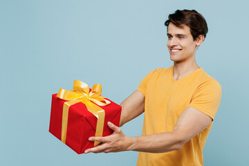 Side view young smiling happy cool man 20s in yellow t-shirt hold giving red present box with gift ribbon bow isolated on plain pastel light blue background studio portrait. People lifestyle concept