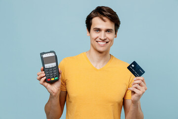 Young smiling happy fun man 20s wear yellow t-shirt hold wireless modern bank payment terminal to process acquire credit card payments isolated on plain pastel light blue background studio portrait