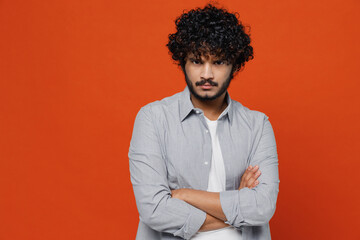 Obraz premium Sullen displeased dissatisfied gloomy young bearded Indian man 20s years old wears blue shirt hold hands crossed isolated on plain orange background studio portrait. People emotions lifestyle concept.