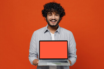 Fascinating charismatic young bearded Indian man 20s years old wear blue shirt hold use work on laptop pc computer with blank screen workspace area isolated on plain orange background studio portrait