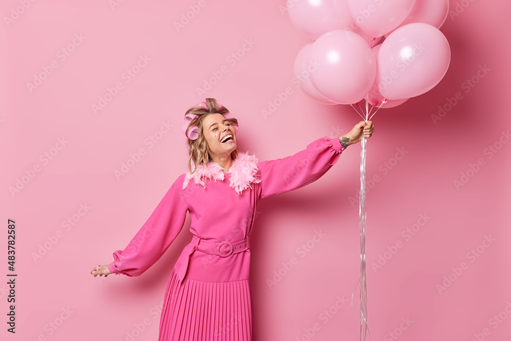 Wall mural Horizontal shot of cheerful European woman weas festive pink dress hair rollers has fun and enjoys celebration dances with bunch of inflated balloons against rosy background has upbeat mood.