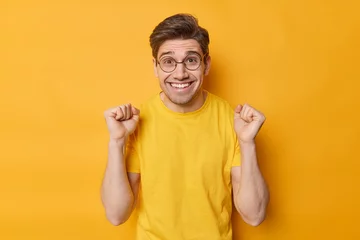 Tuinposter Happy positive man cheers over something clenches fists and rejoices victory smiles toothily feels very glad wears round spectacles and casual t shirt isolated over yellow background. Triumph and luck © Wayhome Studio