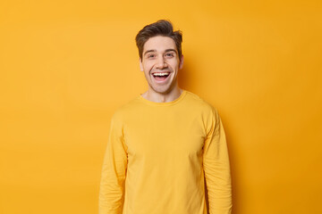 Horizontal shot of happy young European man laughs joyfully expresses positive emotions dressed in casual jumper isolated over vivid yellow background. Monochrome shot. Positive guy indoors.