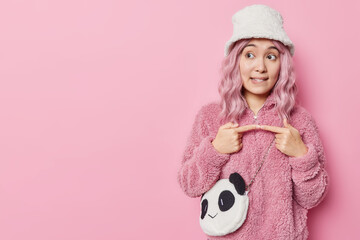 Pensive young Asian woman stands in thoughtful pose tries to decide keeps index fingers opposite each other wears panama and coat isolated over pink background copy space for your promotion.