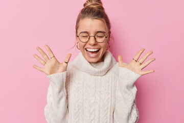 Fototapeta Horizontal shot of happy woman with combed hair raises palms wears round spectacles and white sweater feels amused poses against pink background hears amazing good news screams from happiness obraz