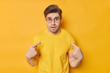 Indignant young man looks puzzled points at himself feels embarrassed wears transparent round spectacles and casual t shirt asks who me isolated over vivid yellow background. Do you blame me
