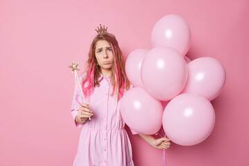 Obraz na płótnie Canvas Unhappy frustrated woman comes on party poses with inflated balloons and magic wand wears small princess crown and dress poses against pink background. People holidays and celebration concept