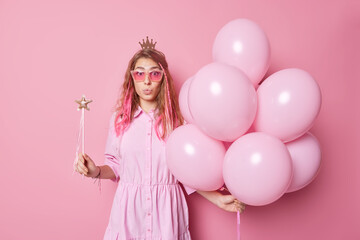 Obraz na płótnie Canvas Impressed astonished fashionable woman wears trendy sunglasses dress and crown on head has image of princess holds bunch of inflated helium balloons magic wand keeps lips rounded poses indoor