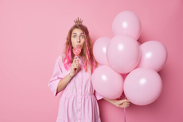 Surprised young woman holds bunch of inflated balloons sweet caramel candy on mouth wears small crown and dress looks with widely opened eyes isolated over pink background. Partying concept.