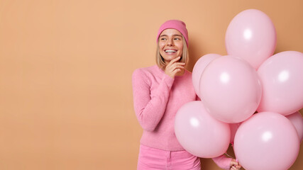 Fototapeta na wymiar Dreamy young lovely woman organizes farewell party touches jawline gently looks away has good mood holds bunch of helium balloons poses against brown background blank copy space for promotion