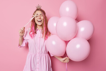 Overjoyed glad young woman poses with bunch of inflated balloons and magic wand has fun on party foolishes around isolated over pink background enjoys holiday. People and celebration concept