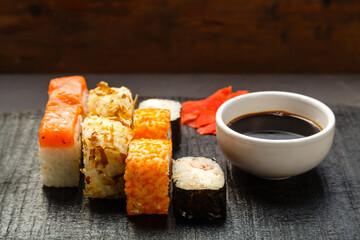 Assorted salmon and shrimp rolls on a black board next to soy sauce and ginger on a dark background.
