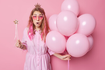 Obraz na płótnie Canvas Impressed stunned European woman wears trendy sunglasses and dress reacts on shocking newsholds bunch of inflated balloons and magic wand isolated over pink background. Holiday event concept